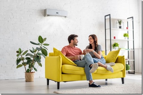 happy young couple talking and holding cups while sitting on yellow sofa under air conditioner at home