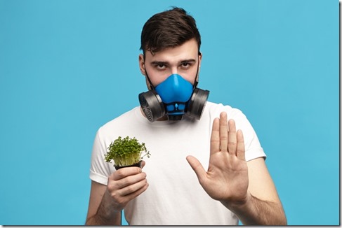 Serious young brunette male in white t-shirt holding micro greens in one hand and making stop gesture with other wearing gas mask, going to spray pesticides to protect plants. Ecology and environment