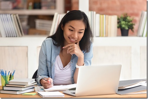 Smiling asian undergraduate teen girl student study in library with laptop books doing online research for coursework, making notes for essay homework assignment, online education e-learning concept