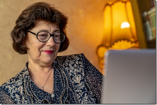 Senior woman in a glasses using laptop. Close up shot. The woman sits on the sofa near to the bookcase. Laptop on the white table. Using technology in old age concept