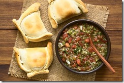 Chilean Pebre sauce, a traditonal condiment made of tomato, onion, garlic, spicy aji pepper and coriander with empanadas on the side, photographed overhead on dark wood with natural light