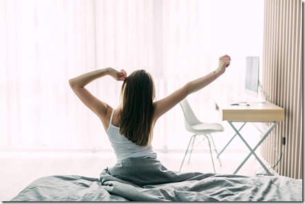 Back view of young woman stretching on unmade bed after waking up and looking at city view in the window. Motivation concept